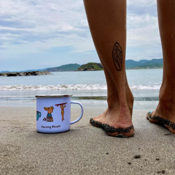 surf mug on the beach next to standing woman with a surfboard tattoo